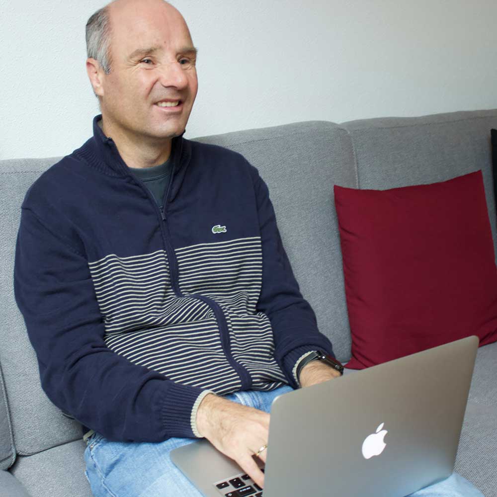 David Röthle schaut lächelnd auf einem Sofa sitzend in die Kamera und hat einen Computer auf dem Schoß..