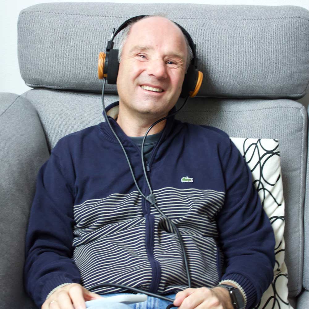 David Röthle schaut lächelnd auf einem Sofa sitzend in die Kamera und hat Kopfhörer auf den Ohren.